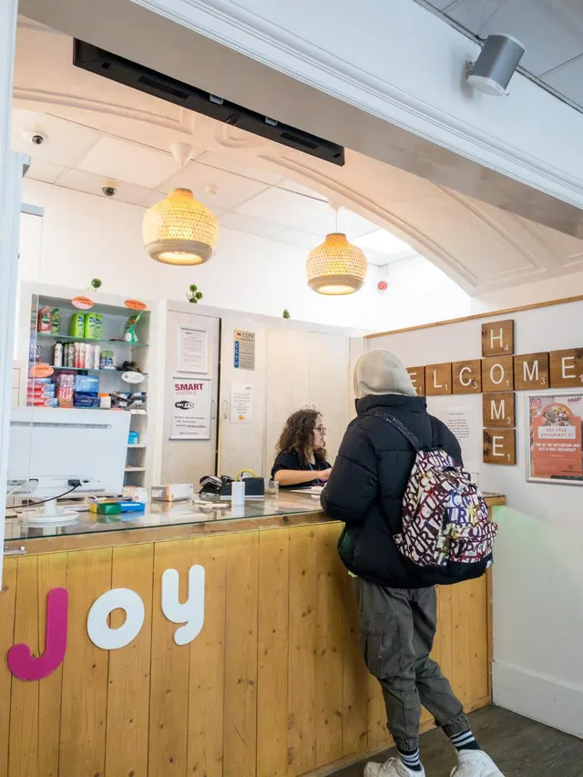 A guest with a backpack stands at a wooden reception desk marked with the letters Joy while a staff member sits behind it.