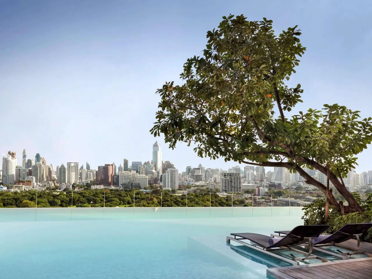 An infinity edge pool with sun loungers overlooks a city skyline of high-rise towers and green trees under a clear sky.
