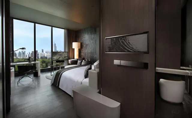A bedroom with a king bed, a desk with a chair and a television, and floor-to-ceiling windows looking out over treetops and a city skyline.