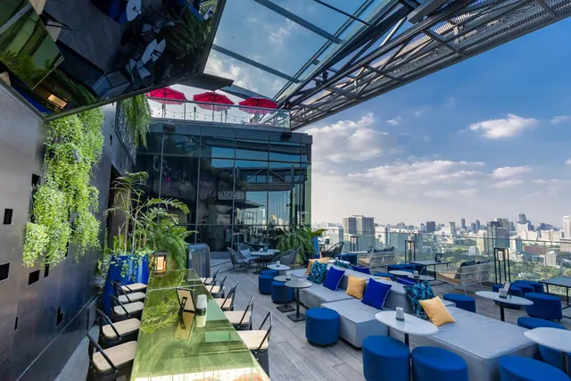 A rooftop bar with blue sofas and low tables, a glass bar counter with chairs, green wall planting, and a city skyline visible beyond the railing.