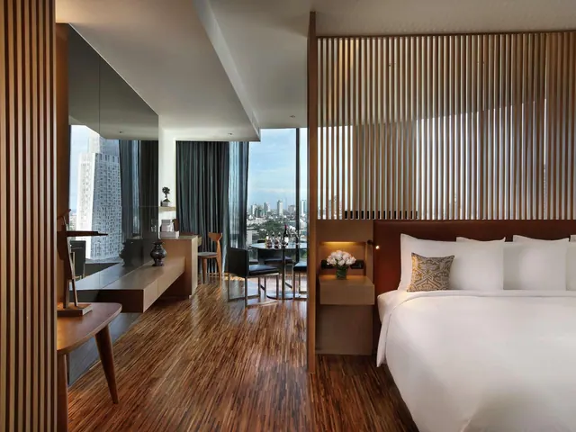A bedroom with a king bed behind a wooden slatted partition, and floor-to-ceiling windows overlooking city towers.