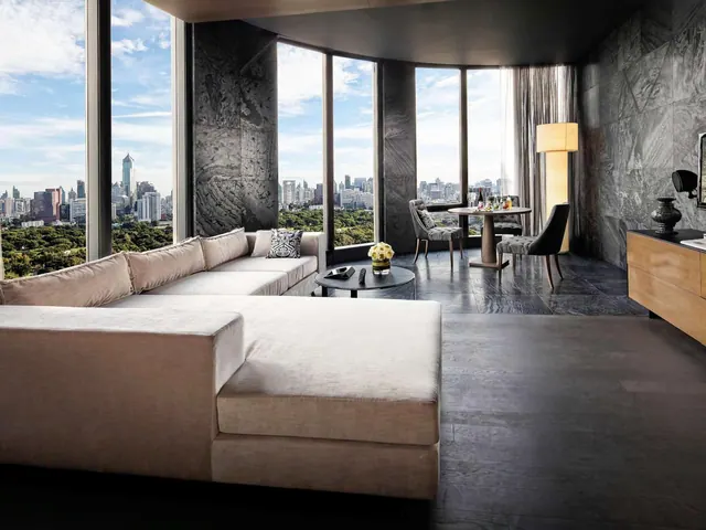 A living area with a large light sectional sofa, a round coffee table, a small dining table by the windows, and a city skyline visible through floor-to-ceiling glass.