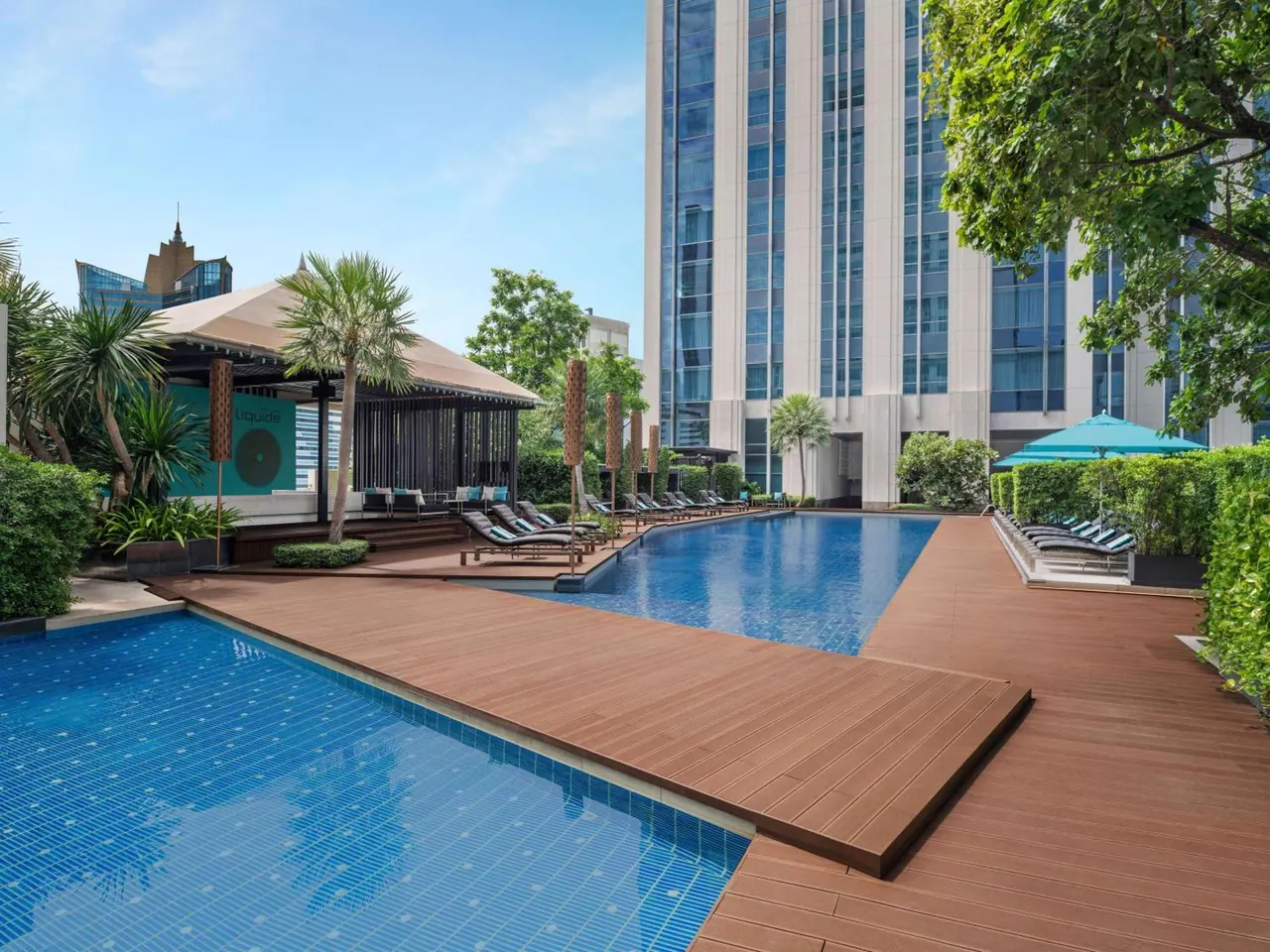 A large blue-tiled pool with a timber deck, sun loungers, teal parasols and a covered pool bar beside a high-rise hotel tower.