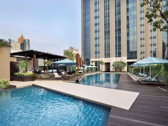 An outdoor pool with blue umbrellas and sun loungers stands beside a tall hotel tower, with a few city buildings in the distance.