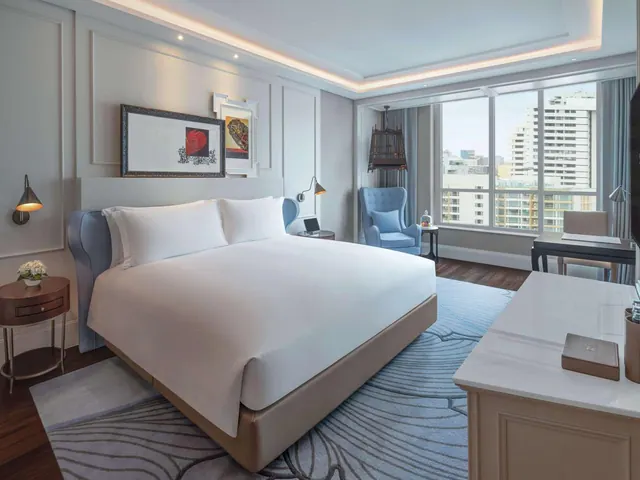 A king bed with white linen stands in a bedroom beside floor-to-ceiling windows that look out over high-rise buildings, with a blue armchair and a desk nearby.