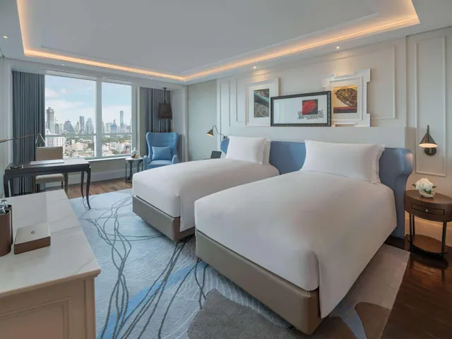 A bedroom with two beds, a desk, a blue armchair, and a city skyline visible through floor-to-ceiling windows.