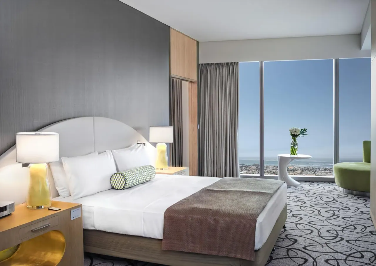 A bedroom with a large white bed, yellow bedside lamps, a green curved armchair and floor-to-ceiling windows looking out over a city and the sea.