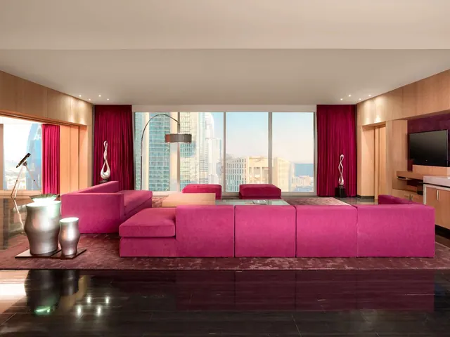 A living room with pink sofas, a wall-mounted television and floor-to-ceiling windows showing city towers outside.