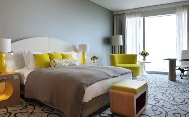 A bedroom with a king bed with yellow cushions, a yellow armchair, a round glass desk with a chair, and floor-to-ceiling windows with sheer curtains.