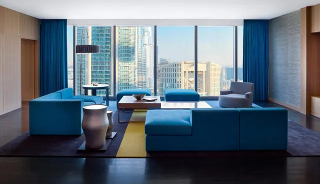A living room with blue sectional sofas, a low wooden coffee table and floor-to-ceiling windows looking out over high-rise towers.