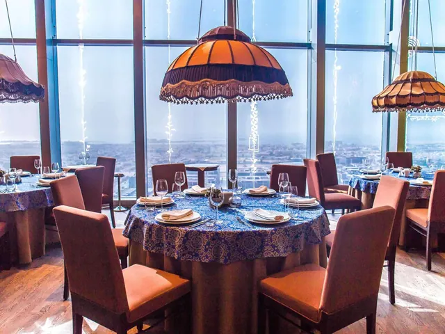 A dining room with round tables set with blue patterned cloths, orange pendant lamps and floor-to-ceiling windows overlooking a hazy city.
