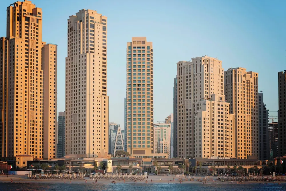 Several tall beige residential towers stand behind a sandy beach with shade umbrellas and a few people in the sea.