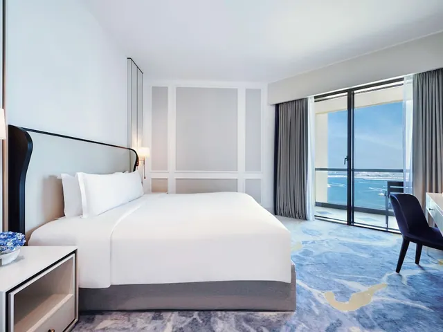 A bedroom with a made king bed, a desk and chair, and sliding glass doors opening onto a balcony with a view of the sea.