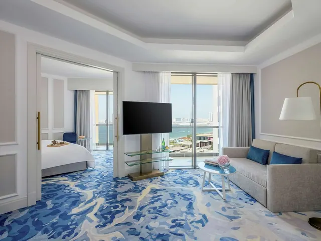 A hotel suite living area with a grey sofa, wall-mounted television and sliding glass doors looking out to the sea, with a bed visible through the open doorway.