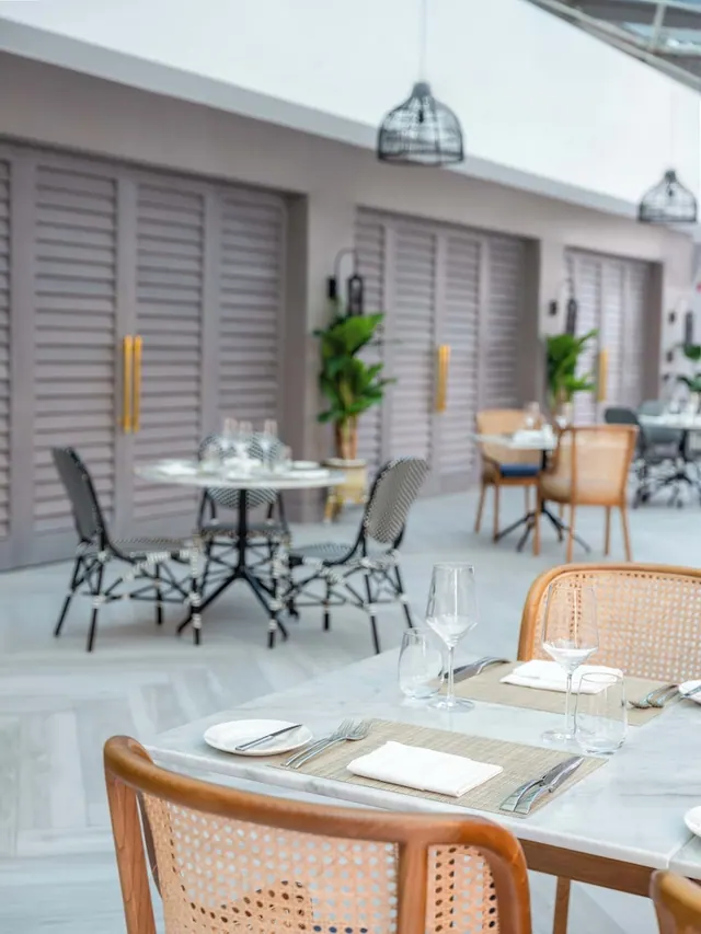 Empty dining area with marble-topped tables, cane chairs, black pendant lights and louvred shutters along the back wall.