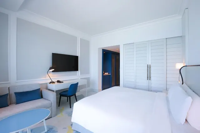 A hotel bedroom with a white bed, a grey sofa with blue cushions, a wall-mounted television, a desk and chair, and louvred wardrobe doors.