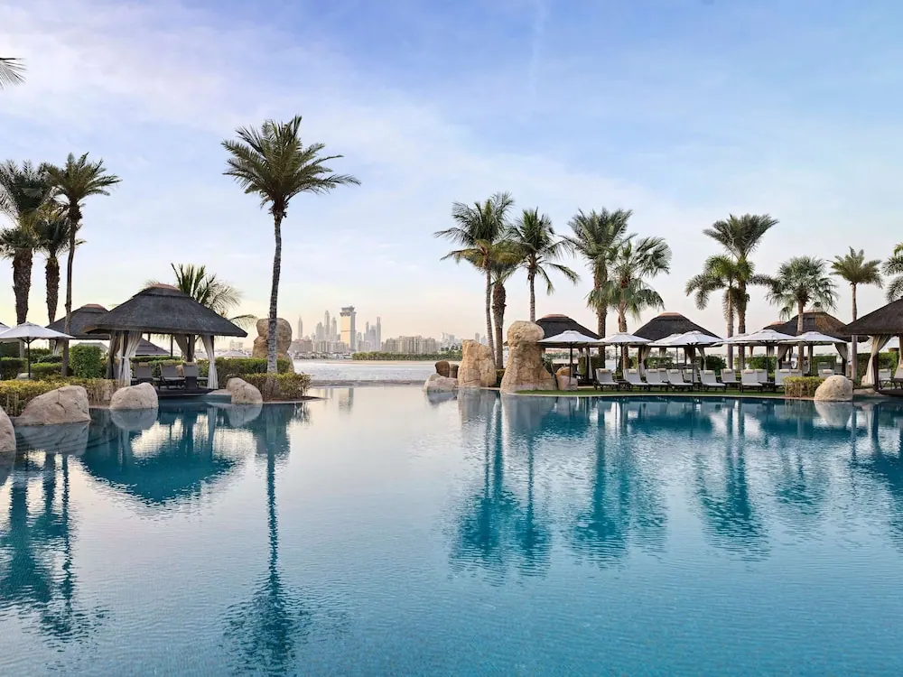 An infinity-edge pool with sun loungers, thatched cabanas and palm trees, with a city skyline visible across the water in the background.