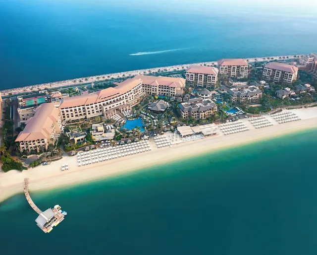 Aerial view of a beachfront resort with a pool, rows of sun loungers and umbrellas on the sand, and a jetty extending over the sea.