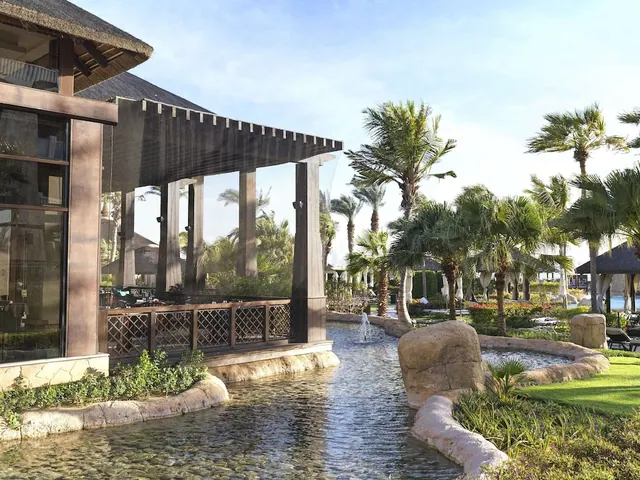 A stone-edged water channel runs past a timber pergola, palm trees and a lawn at a resort, with sun loungers at the right edge.