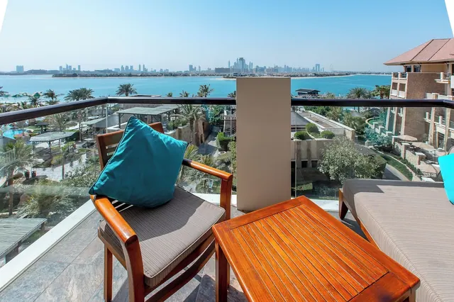 A private balcony with two wooden armchairs, a wooden side table and a daybed, overlooking the sea and a city skyline across the water.