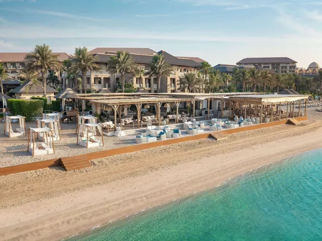 Aerial view of a beachfront lounge with canopied cabanas, pergola-shaded seating and turquoise sea beside a sandy beach, with a hotel and palm trees behind.
