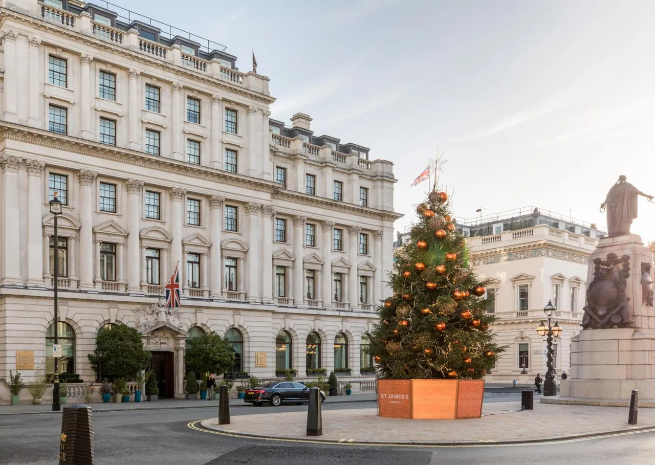 Exterior of a cream stone building with a decorated Christmas tree in an orange planter, a statue on the right and a black car passing on the street.