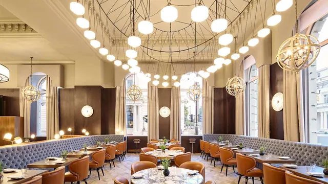 A dining room with marble-topped tables, tan leather chairs, grey tufted banquettes and globe pendant lights, with tall windows showing a stone building outside.