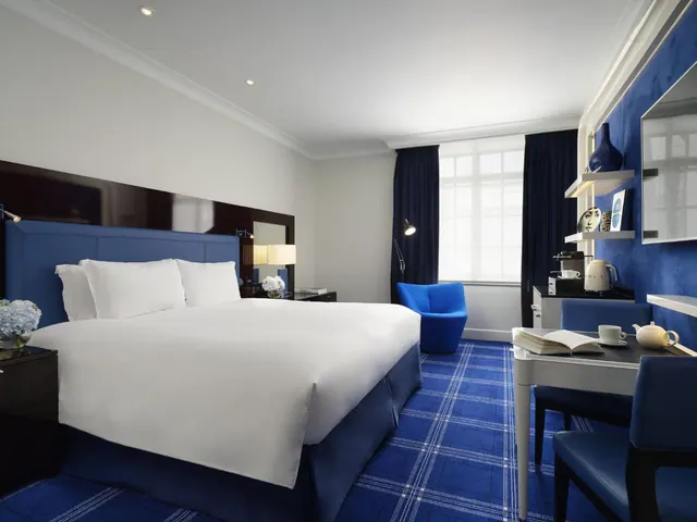 A hotel bedroom with a king bed under white linen, a blue headboard and plaid carpet, a blue armchair by the window, and a desk with a kettle and coffee machine.