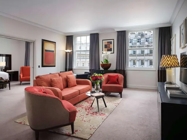 A living room with a coral sofa, two upholstered armchairs, a glass coffee table, and a flat-screen television on a dark sideboard, with tall windows looking out onto a building facade.