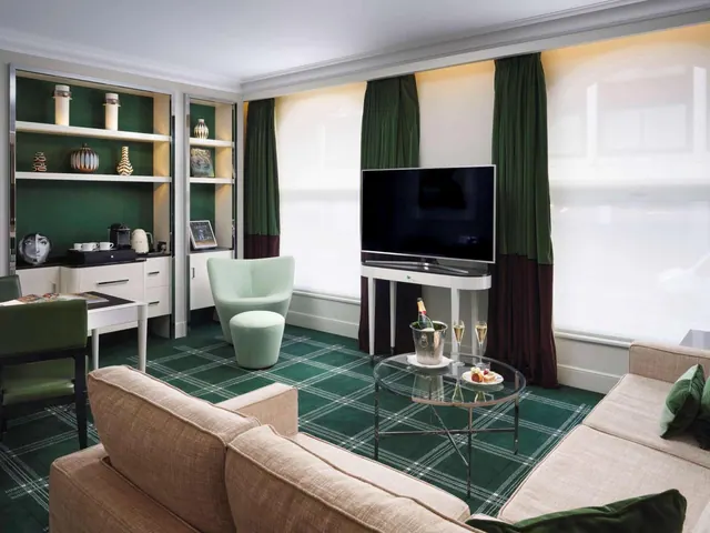 A hotel suite living area with a beige sofa, a wall-mounted television, a glass coffee table holding champagne glasses and a snack plate, and built-in shelving with a kettle and coffee machine on a sideboard.