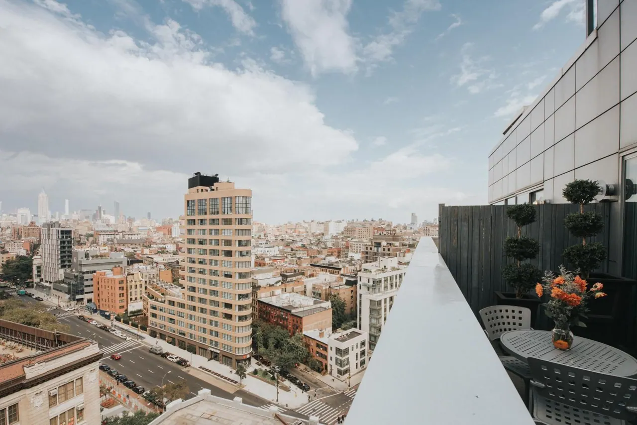 A rooftop terrace with metal chairs and a round table looks out over a city street and dense low-rise buildings, with a tall tower in the middle distance.