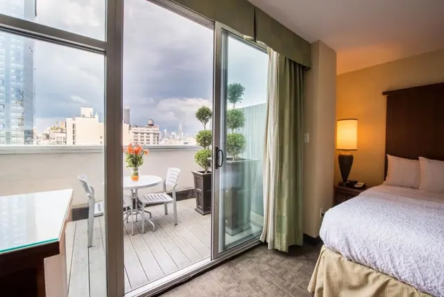 A bedroom with a made bed and sliding glass doors open onto a private balcony with a small table, chairs and potted topiaries, overlooking city buildings and a distant spire.