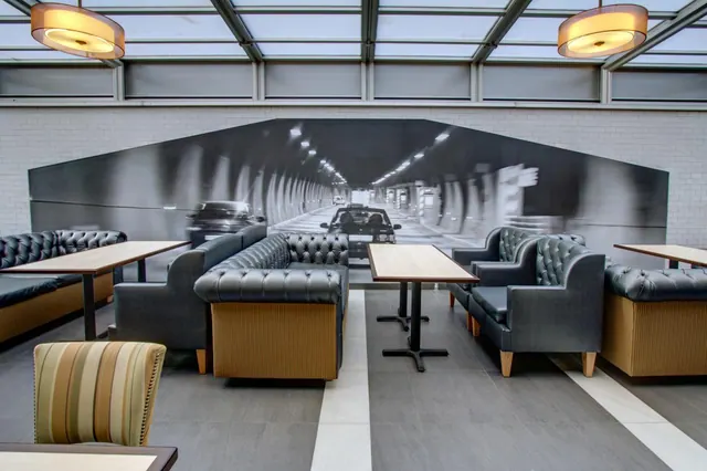 A seating area with dark tufted leather booths, wooden-topped tables and a large black-and-white tunnel mural on the back wall under a glass ceiling.