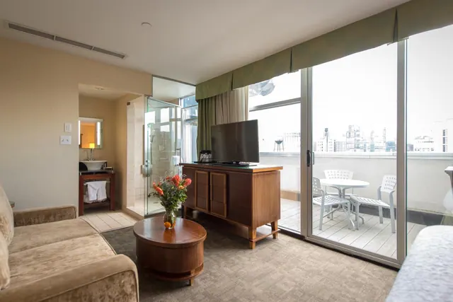 A hotel living area with a sofa, a wooden TV cabinet, a round coffee table with a flower vase, and sliding glass doors onto a balcony with white chairs and a city view.