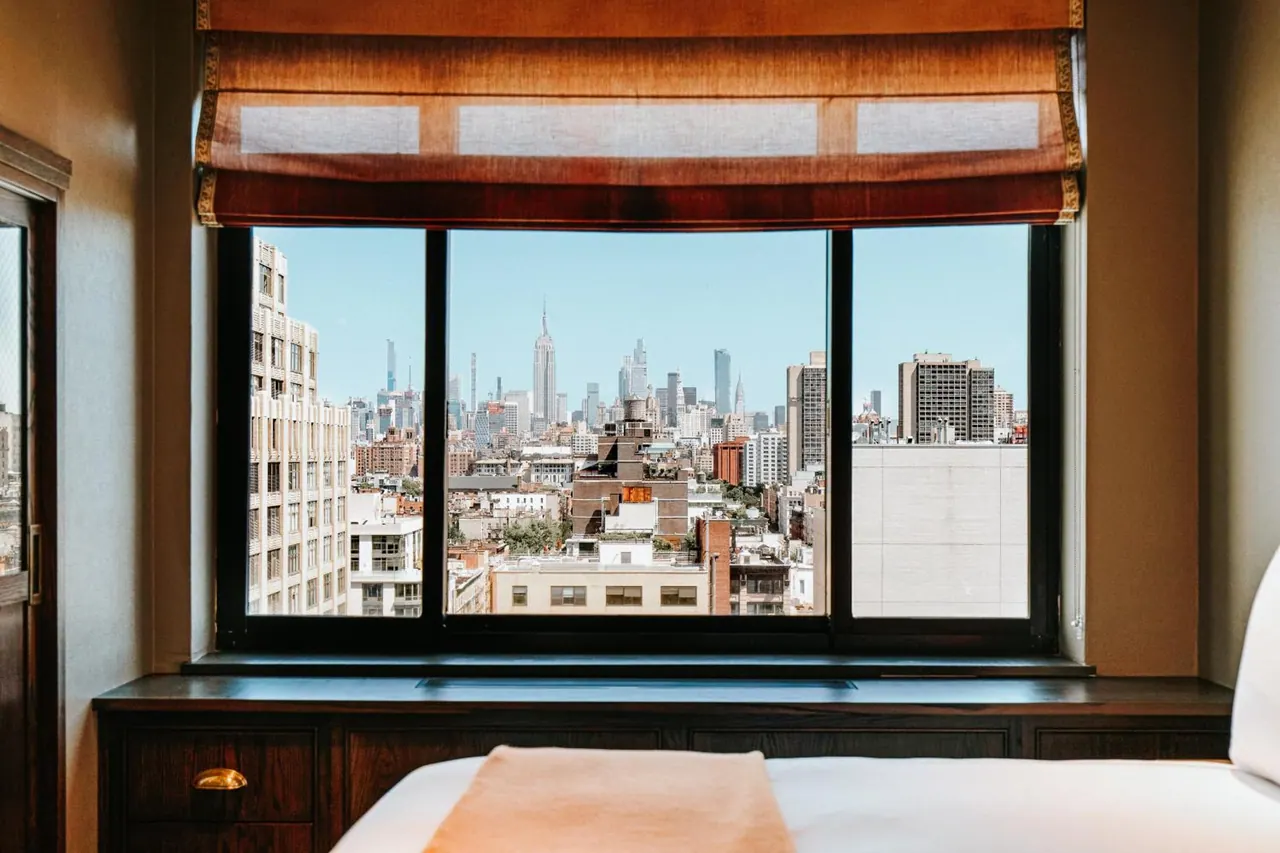 A bedroom window with a tan roman blind looks out over a Manhattan skyline that includes the Empire State Building, with a white bed in the foreground.