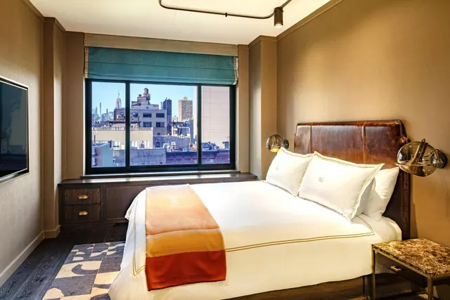 A king bed with a leather headboard and white bedding sits beside a window showing city buildings, including the Empire State Building.