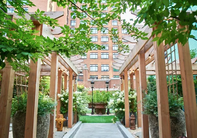 A covered wooden pergola walkway leads toward a lawn and a stone fountain in a courtyard enclosed by a brick building with many windows.