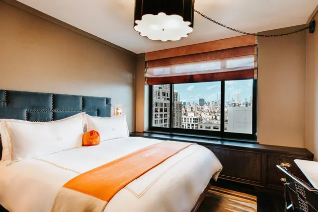A bedroom with a king bed, a dark upholstered headboard, a wooden cabinet under the window, and a view of city buildings through the window.