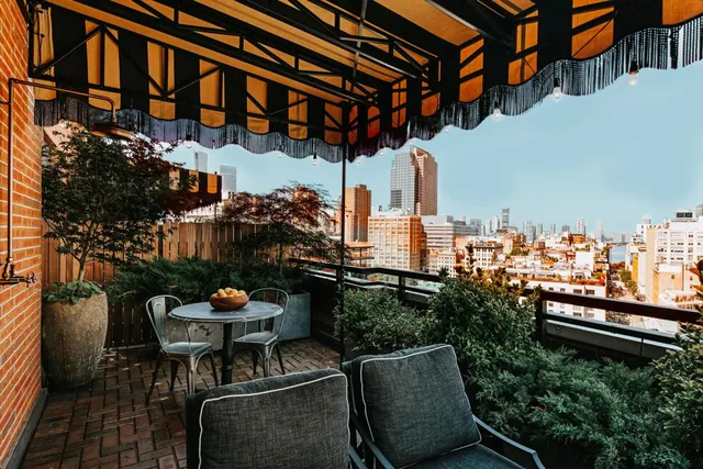 A brick-floored rooftop terrace with a striped awning, a metal table and chairs, grey lounge seats, potted plants and a view of city high-rise buildings.