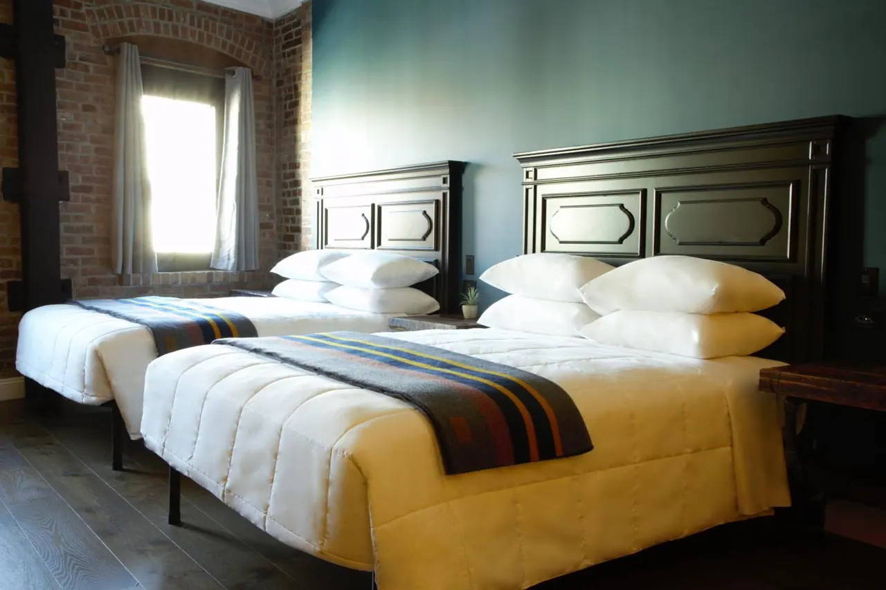 Two beds with white bedding, dark wooden headboards and striped throws stand in a room with exposed brick walls and a teal wall, beside a window with white curtains.