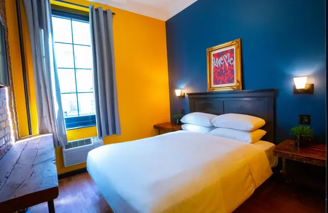 A bedroom with a double bed, yellow and dark blue walls, a framed red artwork above the headboard, and a wall air conditioner under the window.