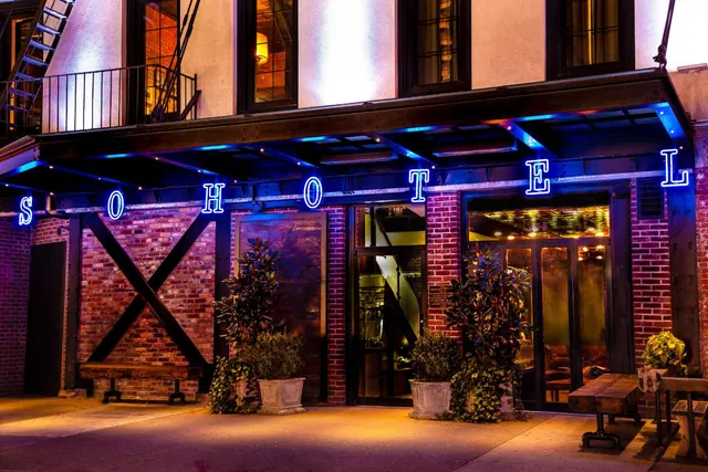 Night exterior of a brick building with blue neon letters spelling SOHOTEL above the entrance, flanked by potted plants and a wooden bench.