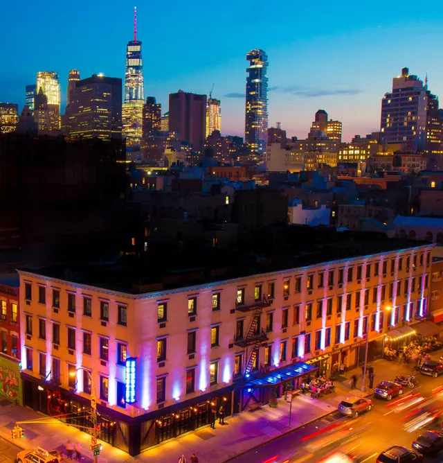 Twilight elevated view of a lit building with purple uplighting, street traffic below, and the Lower Manhattan skyline behind, including One World Trade Center.
