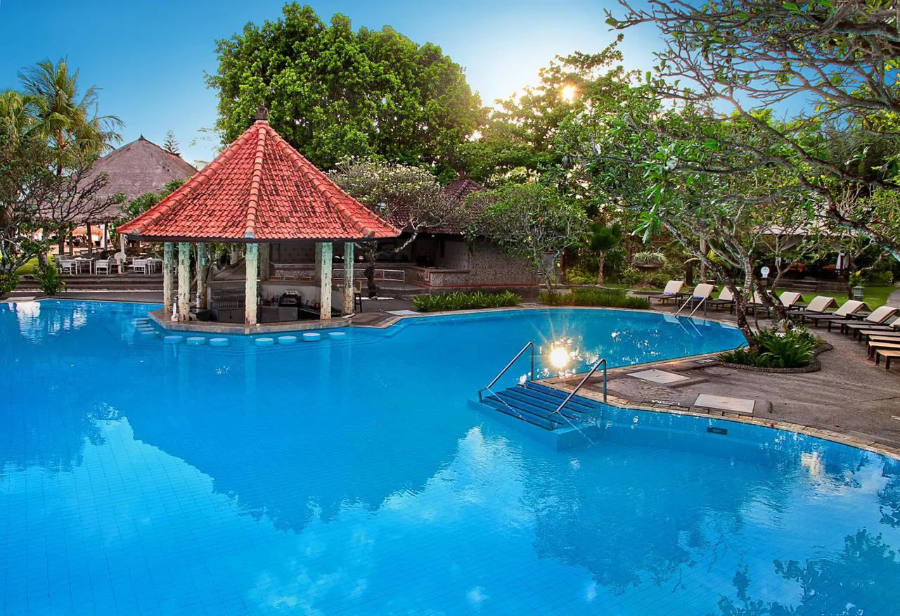 A large blue swimming pool with a red-tiled open pavilion, sun loungers and tall trees around it in low evening sunlight.