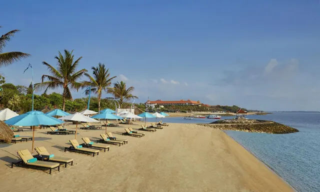A sandy beach with rows of sun loungers under light blue umbrellas, palm trees, a rocky breakwater, and the sea on the right, with a red-roofed building in the distance.