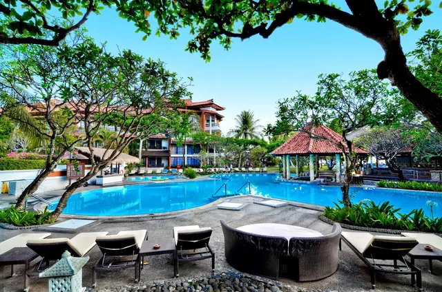 A large outdoor swimming pool with sun loungers and a round wicker table in the foreground, with a tiled gazebo and multi-storey hotel buildings beyond the trees.