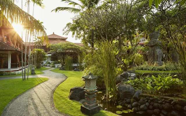 A stone path curves past a lawn, a stone lantern, a statue and a small lily pond beside traditional Balinese buildings with red-tiled roofs.