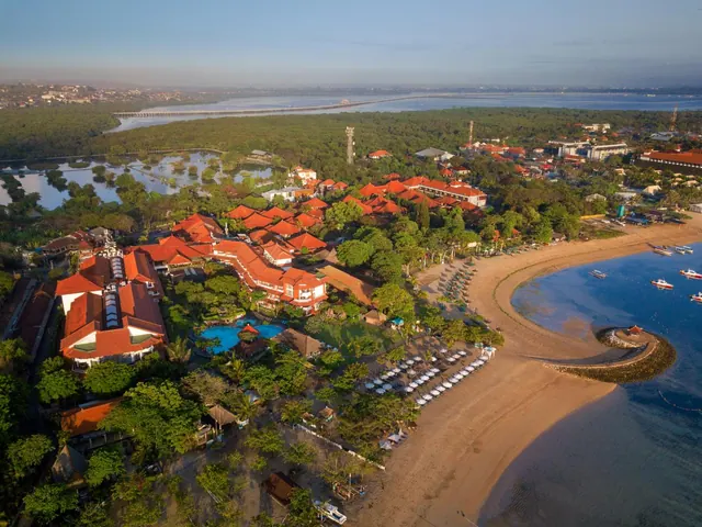 Aerial view of a beachfront resort with red-roofed buildings, a swimming pool, rows of sun loungers and umbrellas on the sand, and the sea to the right.
