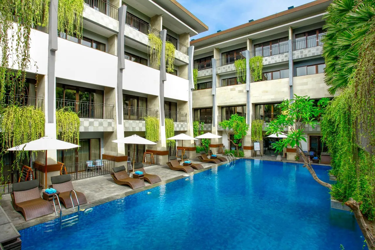 A large blue swimming pool is surrounded by sun loungers and parasol umbrellas, with multi-storey hotel buildings and hanging greenery around the courtyard.