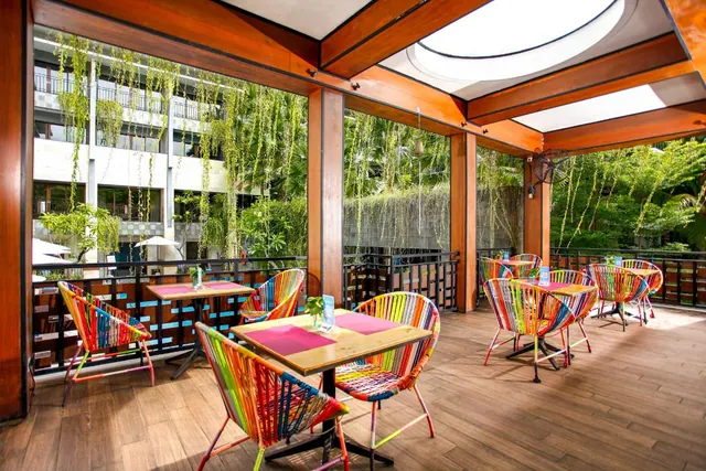 A covered outdoor terrace with colorful woven chairs and wooden tables, with a view past glass panels onto green plants and a hotel building across a courtyard.